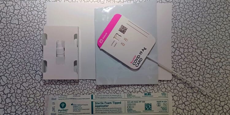 The image shows components of a COVID-19 rapid test kit. It includes a test card labeled "COVID-19 Ag CARD" with a swab attached, a small bottle in a white holder, and a sterile foam-tipped applicator in its packaging. The items are placed on a surface with a cracked pattern.