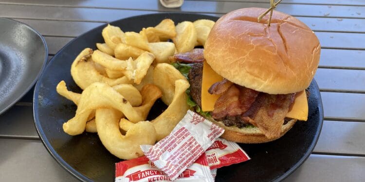 A plate with a cheeseburger and curly fries. The burger includes bacon, cheese, and lettuce, and is held together with a skewer. There are packets of ketchup on the plate. In the background, there is a small plate with rice and a rolled napkin on a table.