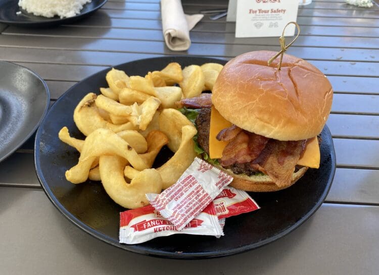 A plate with a cheeseburger and curly fries. The burger includes bacon, cheese, and lettuce, and is held together with a skewer. There are packets of ketchup on the plate. In the background, there is a small plate with rice and a rolled napkin on a table.