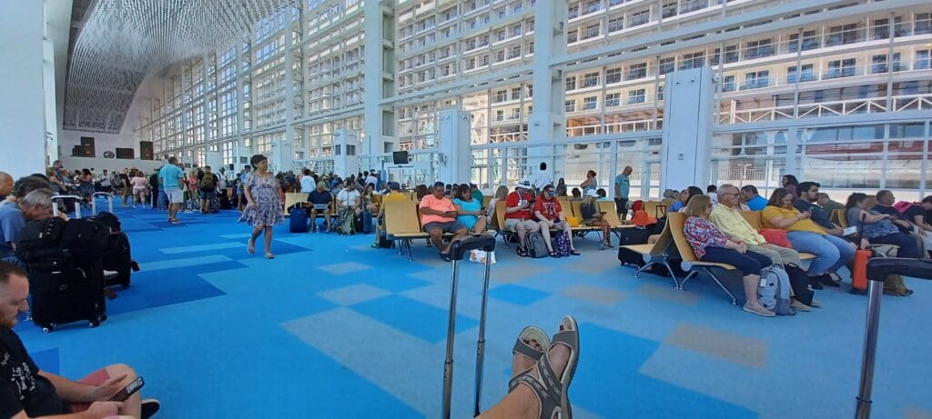 The image shows a large, open waiting area with many people seated on rows of chairs. The floor is covered with a blue carpet, and the space is enclosed by tall glass walls, allowing natural light to fill the area. The ceiling is high and features a modern design. Some people are standing or walking, while others are sitting with luggage nearby. The atmosphere appears to be that of a busy terminal or waiting lounge.