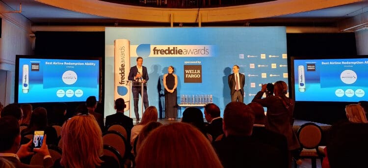 The image shows an awards ceremony for the Freddie Awards, sponsored by Wells Fargo. A man is speaking at a podium on stage, with a woman standing beside him. Another man stands to the right. The backdrop displays the Freddie Awards logo and sponsor information. Two screens on either side of the stage show the category "Best Airline Redemption Ability" for the Americas, with "AAdvantage" highlighted. The audience is seated, and some people are taking photos.
