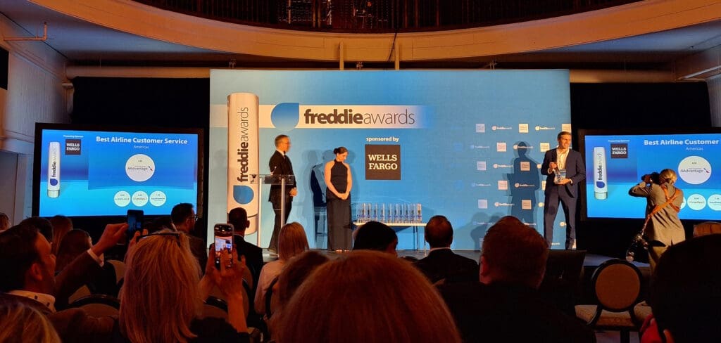 The image shows an awards ceremony stage with a blue backdrop featuring the "freddie awards" logo and a "sponsored by WELLS FARGO" sign. Two people are standing on stage, one holding an award. There are two screens displaying "Best Airline Customer Service Americas" with a chart showing "AAdvantage" and other airline programs. Audience members are seated, some taking photos.