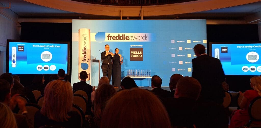 The image shows an awards ceremony with a stage and a large screen displaying "Best Loyalty Credit Card Americas" and the sponsor "Wells Fargo." Two people are standing on stage, one holding an award. The backdrop features the "freddie awards" logo. The audience is seated in front of the stage, and several trophies are visible on a table.