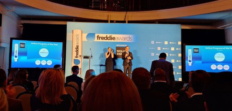 The image shows an awards ceremony stage with a large blue backdrop displaying the "freddie awards" logo and "sponsored by Wells Fargo." Two people are standing on stage, clapping. In the foreground, there is an audience seated and watching. Two screens on either side of the stage display the text "Airline Program of the Year, Americas," with "Aeroplan" highlighted as the winner. The event appears to be formal, with attendees dressed in business attire.