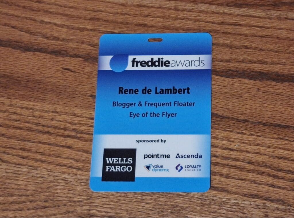 The image shows a blue name badge for the "freddie awards." It has the name "Rene de Lambert" with the title "Blogger & Frequent Floater" and "Eye of the Flyer" written below. The badge is sponsored by several companies, including Wells Fargo, point.me, Ascenda, value dynamx, and Loyalty Status Co. The badge is placed on a wooden surface.
