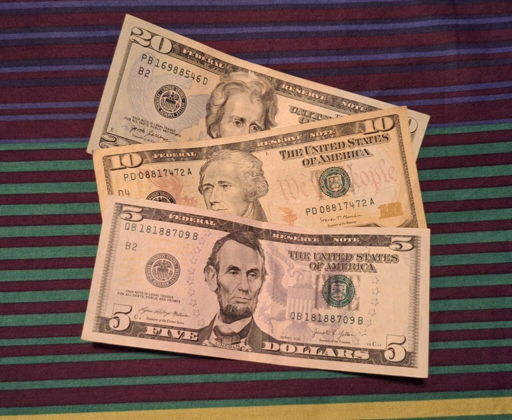 The image shows three U.S. dollar bills placed on a striped fabric background. The bills are a $20 bill, a $10 bill, and a $5 bill, each featuring portraits of historical figures. The fabric has horizontal stripes in shades of purple, green, and brown.