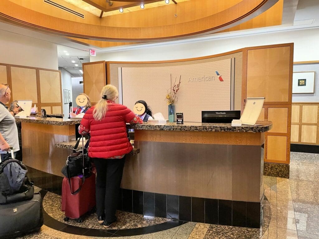 A woman in a red jacket is standing at a reception desk in an airport lounge, speaking with two staff members. The desk has a granite countertop and a sign with the American Airlines logo. There are suitcases next to the woman, and the area has a modern design with wood paneling and marble flooring.