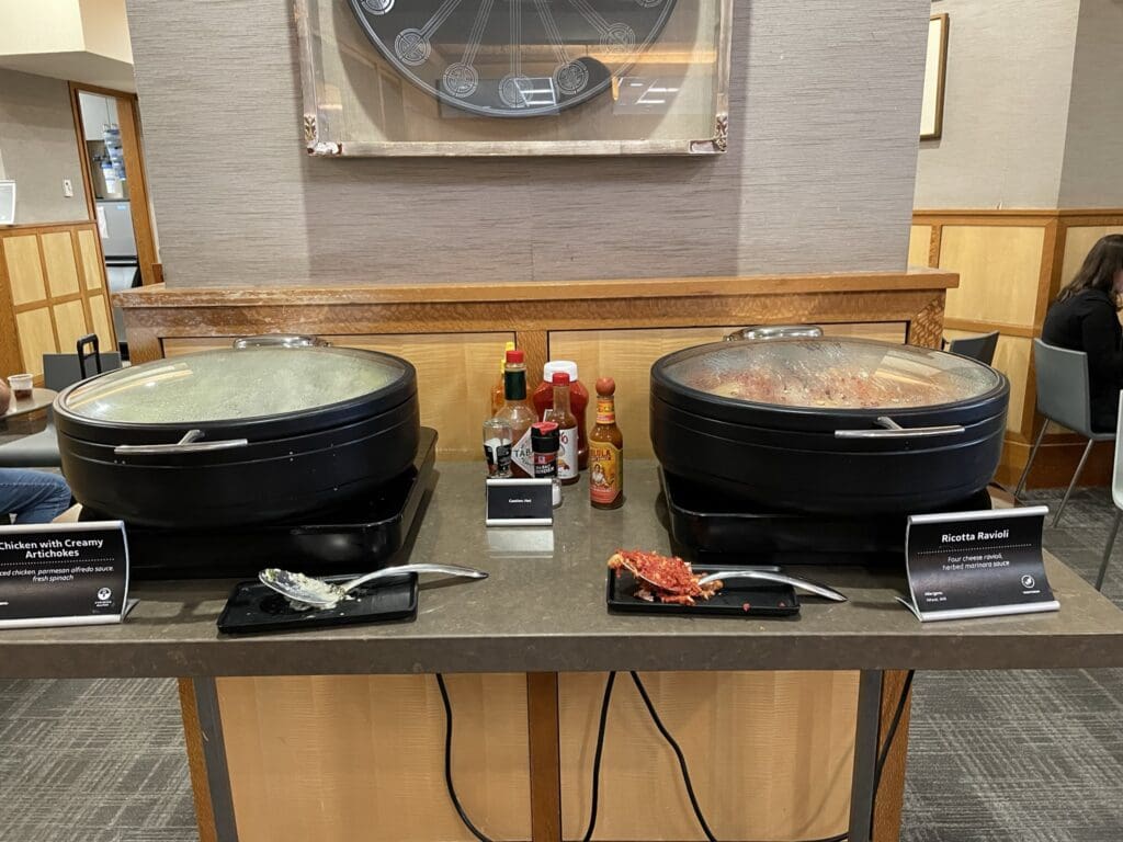 The image shows a buffet setup with two covered serving dishes on a counter. The left dish is labeled "Chicken with Creamy Artichokes," and the right dish is labeled "Ricotta Ravioli." In between the dishes, there are various condiments, including hot sauce and ketchup. A serving spoon with some food residue is placed in front of each dish. The background features a wall with wood paneling and a large decorative clock.