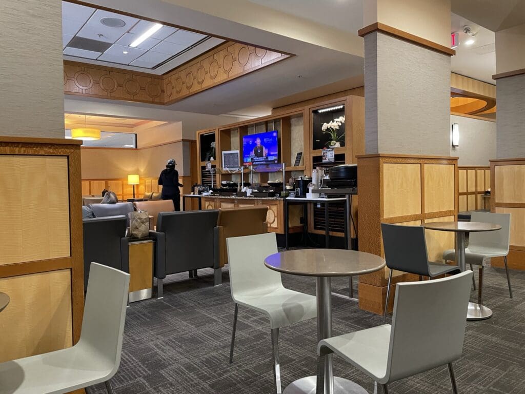 The image shows a lounge area with modern decor. There are several round tables with gray chairs in the foreground. In the background, there are gray cushioned chairs and a small counter with a television mounted above it. A person is standing near the counter, and another person is seated on a chair. The lighting is warm, with ceiling lights and a lamp providing illumination. The walls and columns are decorated with light wood paneling.