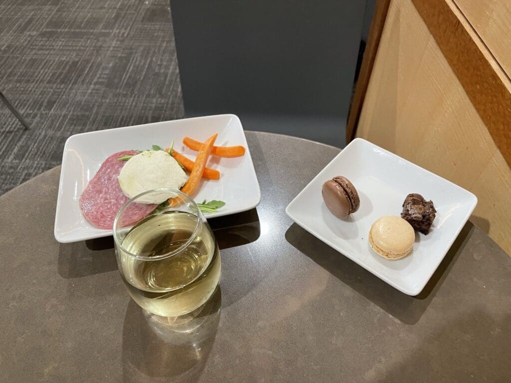 A table with two white plates and a glass of white wine. The left plate contains slices of salami, a scoop of cheese, and carrot sticks. The right plate has a chocolate macaron, a vanilla macaron, and a small piece of chocolate brownie.