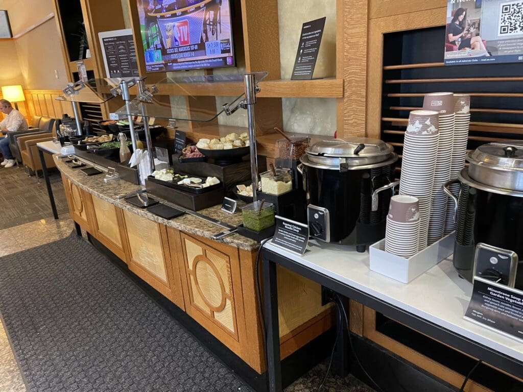 The image shows a buffet setup in a lounge area. There are various food items displayed on a long counter, including bowls of bread, containers with toppings, and two large soup pots. Stacks of disposable cups are placed next to the soup pots. Above the counter, a television is mounted on the wall, showing a sports channel. In the background, a person is seated on a couch. The area has a warm, inviting atmosphere with wooden paneling and soft lighting.