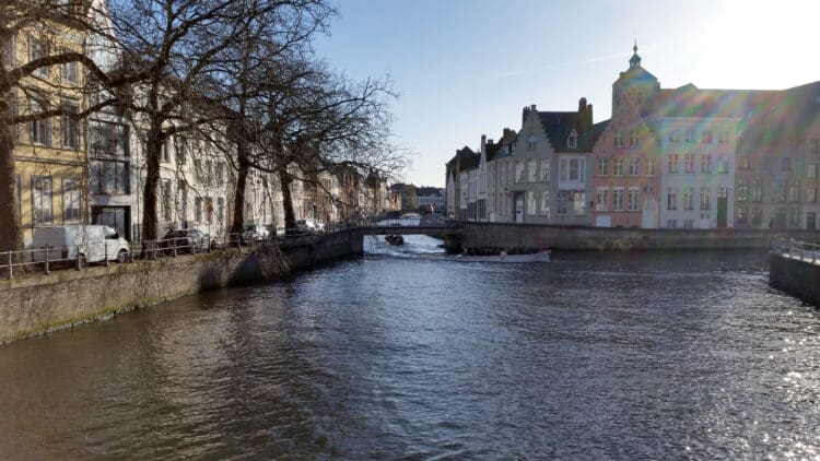 A scenic canal view in Bruges, Belgium, with historic buildings lining both sides. A boat with passengers is traveling along the canal under a small bridge. Leafless trees are visible along the left side, and the sun is shining brightly, creating a lens flare effect.