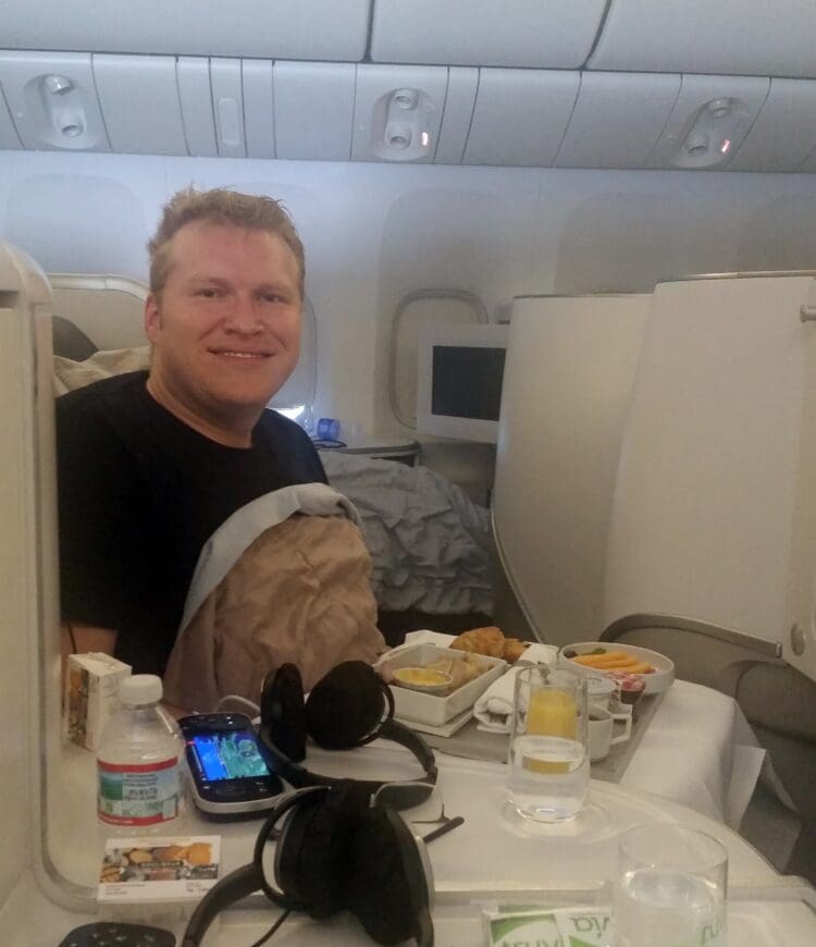 A person is sitting in a business class airplane seat, smiling at the camera. In front of them is a tray with a meal, including a croissant, fruit, and a drink. There are headphones, a bottle of water, and a handheld gaming device on the table. The cabin interior is visible in the background.
