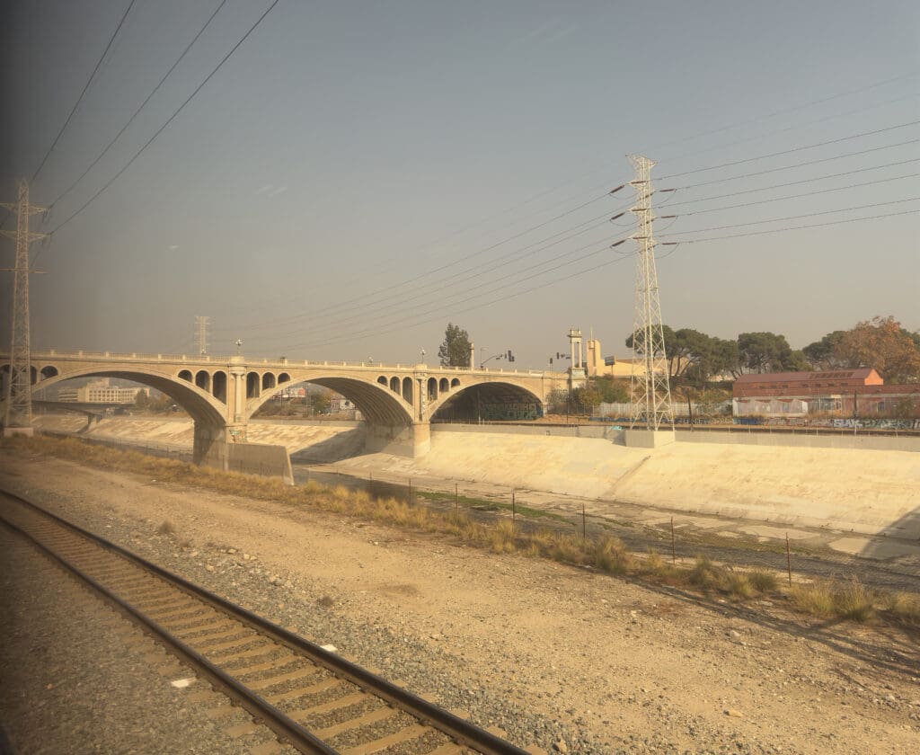 A concrete bridge with multiple arches spans over a dry riverbed or canal. In the foreground, there are train tracks running parallel to the riverbed. Power lines and a transmission tower are visible, and there are buildings and trees in the background under a clear sky.