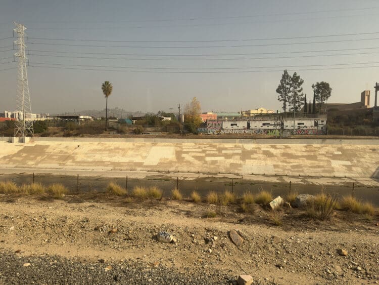 The image shows a dry concrete riverbed with a small amount of water running through it. In the background, there are industrial buildings and structures, including a power line tower. Graffiti is visible on a wall along the riverbank. A few trees, including a tall palm tree, are scattered throughout the scene. The foreground features dry, rocky terrain with sparse vegetation. The sky is clear and slightly hazy.