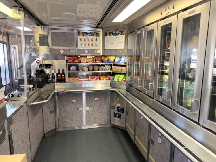 The image shows the interior of a train's snack bar or café area. There are shelves stocked with various snacks, including chips, candy, and packaged goods. A coffee machine and bottles of beverages are visible on the counter. The area is enclosed with stainless steel cabinets and glass-fronted refrigerators. Signs and menus are displayed above the shelves.
