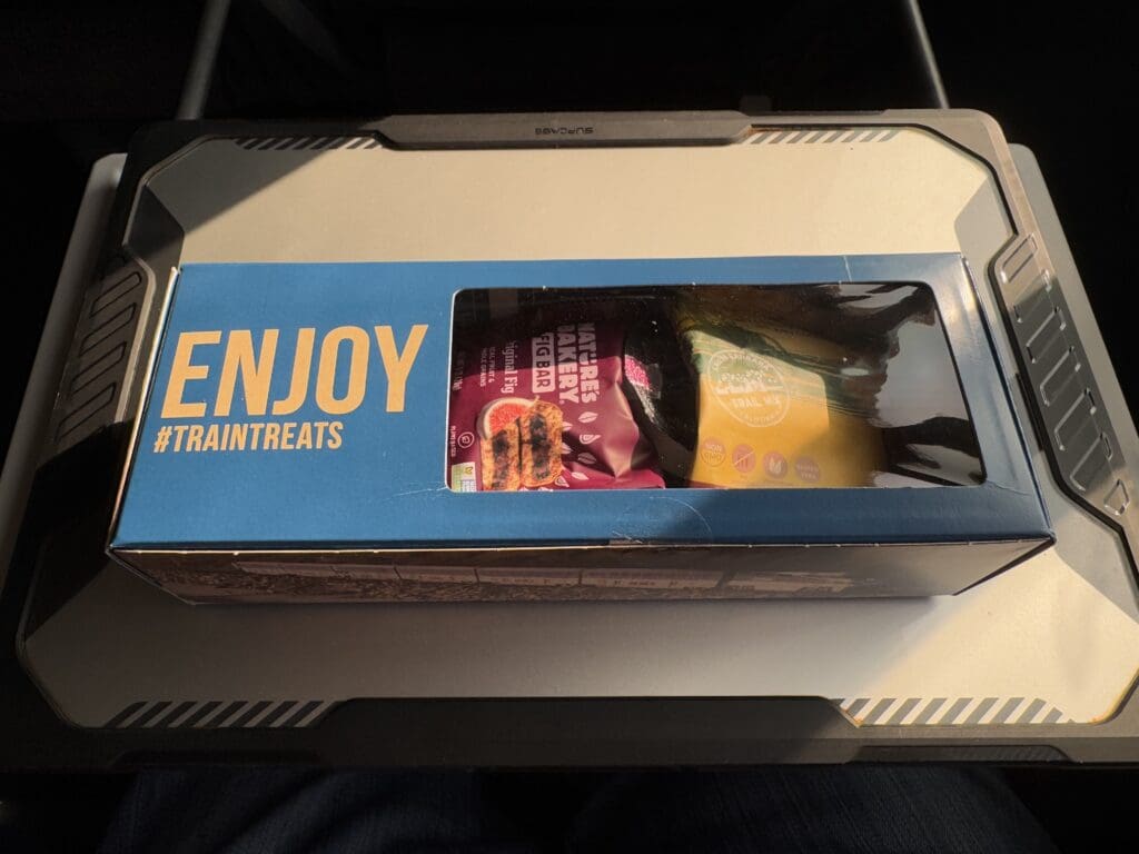 The image shows a box with a blue lid featuring the words "ENJOY #TRAINTREATS" in large yellow letters. The box has a transparent window through which packaged snacks are visible, including a fig bar and a trail mix. The box is placed on a tray table, likely on a train.