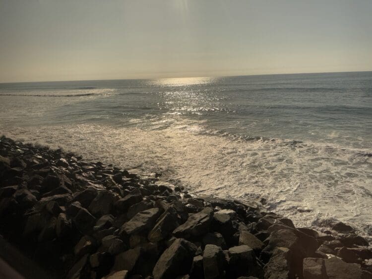 A serene coastal scene with sunlight reflecting off the ocean. Waves gently crash onto a rocky shoreline in the foreground, while the horizon stretches out under a clear sky.