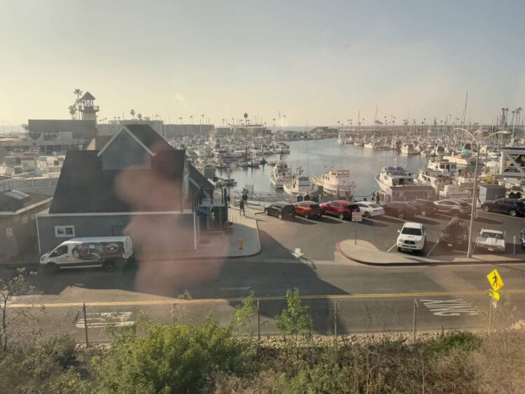 The image shows a marina with numerous boats docked in the water. In the foreground, there is a building with a dark roof and a white van parked nearby. Several cars are parked along the street, and a pedestrian crossing sign is visible. The background features a clear sky and palm trees lining the horizon.