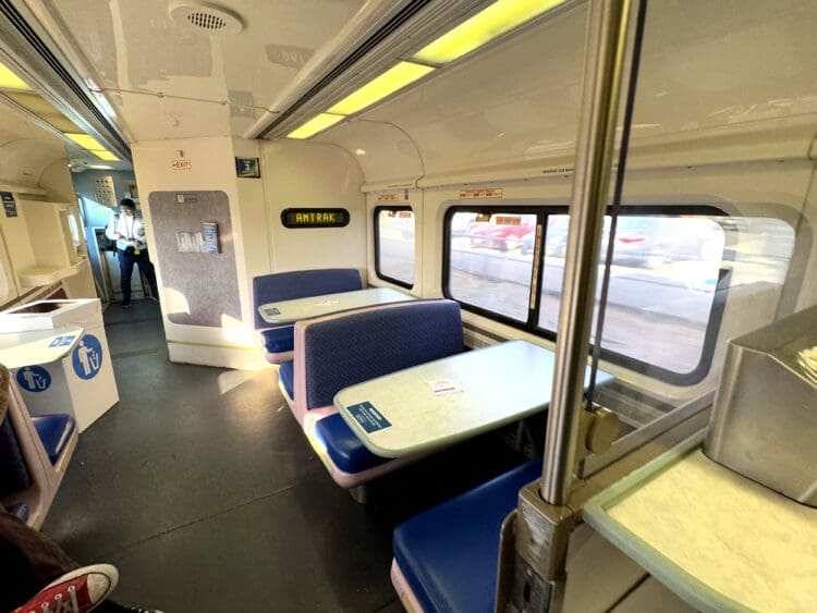 The image shows the interior of an Amtrak train car. There are blue cushioned booth-style seats with tables on the right side. A person is standing near the entrance at the back of the car. The windows on the right side show a blurred view of the outside. Overhead lighting illuminates the space, and there are signs and a trash receptacle visible.