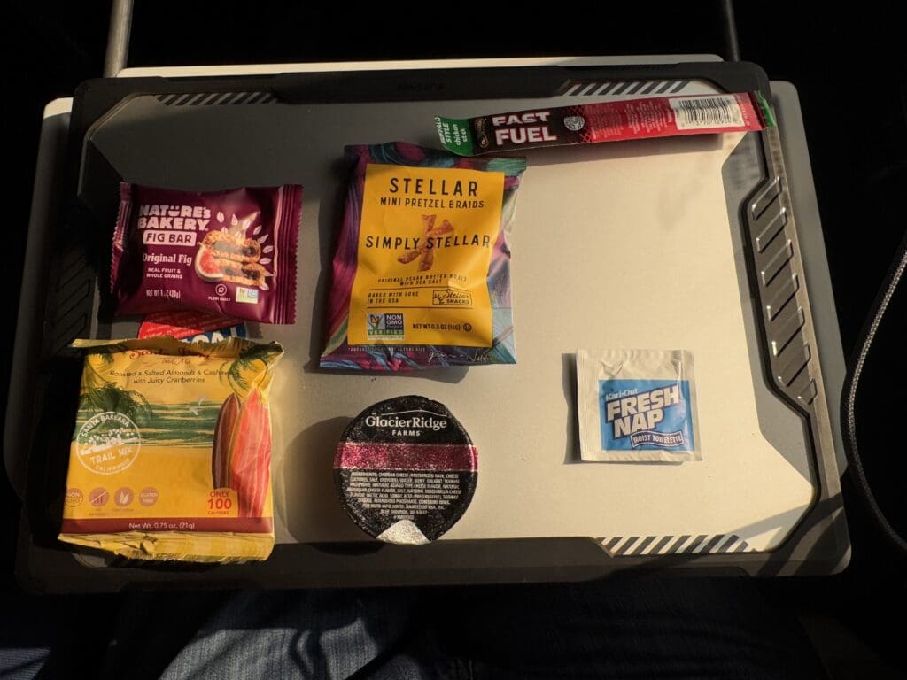 The image shows a collection of snacks and a moist towelette arranged on a tray. The items include:1. A Nature's Bakery Fig Bar in Original Fig flavor. 2. A Stellar Mini Pretzel Braids package. 3. A Fast Fuel energy stick. 4. A packet of trail mix with roasted and salted almonds, cashews, and cranberries. 5. A cup labeled Glacier Ridge Farms. 6. A Fresh Nap moist towelette. The tray is placed on someone's lap, and the lighting suggests it is taken in a well-lit environment.