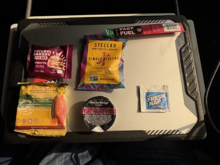 The image shows a collection of snacks and a moist towelette arranged on a tray. The items include:

1. A Nature's Bakery Fig Bar in Original Fig flavor.
2. A Stellar Mini Pretzel Braids package.
3. A Fast Fuel energy stick.
4. A packet of trail mix with roasted and salted almonds, cashews, and cranberries.
5. A cup labeled Glacier Ridge Farms.
6. A Fresh Nap moist towelette. 

The tray is placed on someone's lap, and the lighting suggests it is taken in a well-lit environment.