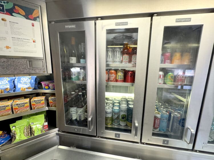 The image shows a section of a convenience store or cafe with three glass-door refrigerators. The left refrigerator contains bottles of wine and other beverages. The middle refrigerator has cans of various drinks, including energy drinks and iced tea. The right refrigerator contains more canned beverages, including soda. To the left of the refrigerators, there are shelves with snacks such as chips, instant noodles, and pistachios. A menu is visible on the wall above the snacks.