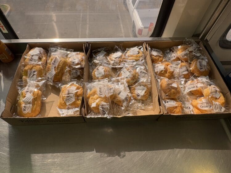 The image shows a cardboard box containing individually wrapped pastries. The pastries are arranged in rows, and each is sealed in clear plastic packaging with a label. The box is placed on a metal surface, and there is a window in the background through which a parked vehicle is visible.