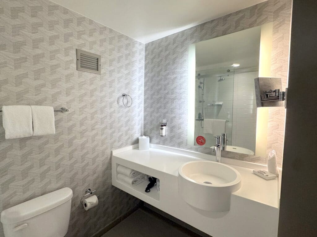 The image shows a modern bathroom with a geometric patterned wallpaper. There is a white toilet with a roll of toilet paper on a holder. Above the toilet, a towel rack holds two white towels. The sink area features a round basin with a sleek faucet, set in a white countertop. A large, illuminated mirror is mounted on the wall above the sink. Below the countertop, there are additional towels and a hairdryer. The shower area is visible in the reflection of the mirror, with a glass door and white tiles.