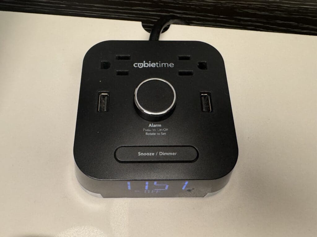 The image shows a black digital alarm clock with the brand name "cubie time" on top. It features two USB ports, a central knob for setting the alarm, and a button labeled "Snooze / Dimmer." The digital display at the bottom shows the time "11:51." The clock is placed on a light-colored surface.