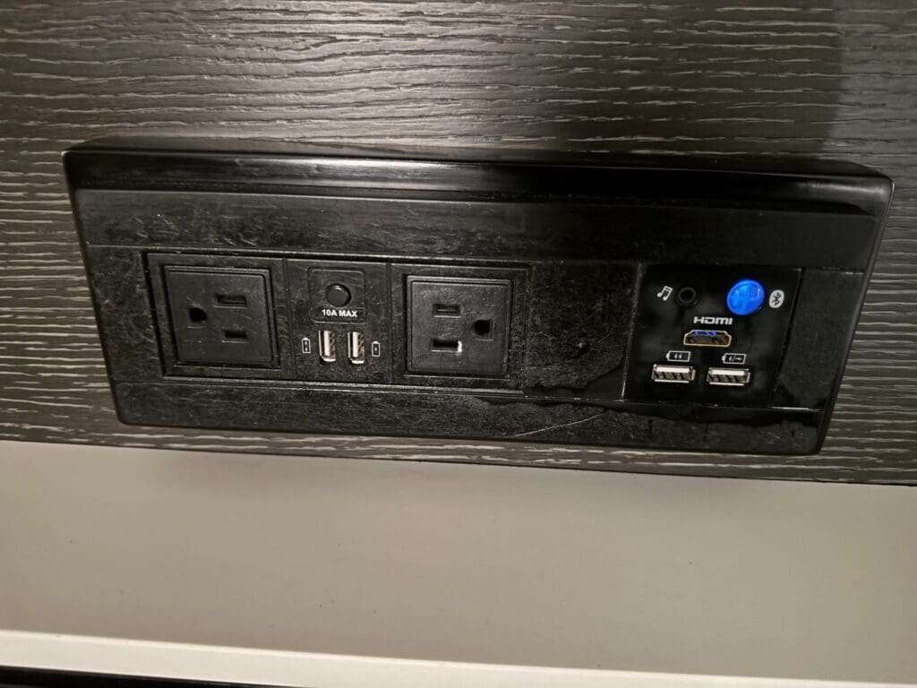 The image shows a black wall-mounted panel with various electrical and connectivity outlets. It includes two standard power outlets, two USB ports labeled "10A MAX," an HDMI port, and two additional USB ports. There is also a blue button with a Bluetooth symbol and a music note icon. The panel is set against a dark wood-textured background.