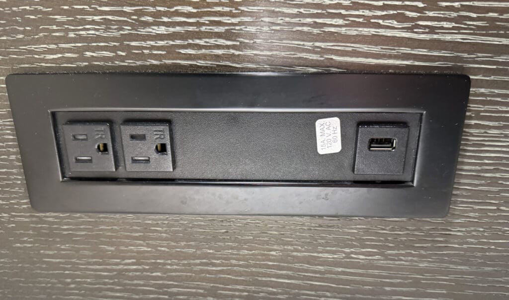 The image shows a black electrical outlet panel mounted on a wooden surface. The panel includes two standard three-prong electrical sockets and one USB port. There is a label next to the USB port indicating "15A MAX, 120V AC, 60Hz."