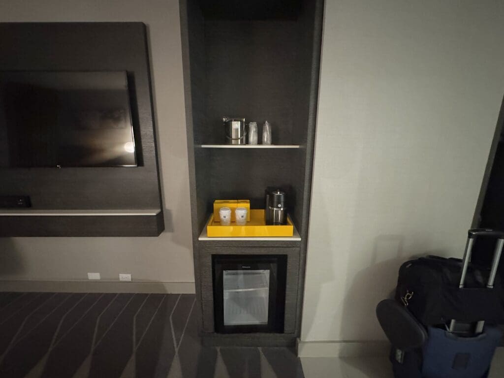 The image shows a hotel room setup with a wall-mounted flat-screen TV on the left. To the right, there is a built-in shelf unit. The top shelf holds an ice bucket and glasses, while the middle shelf has a yellow tray with two disposable coffee cups and a coffee maker. Below, there is a small refrigerator. A piece of luggage with a handle is visible on the floor to the right.