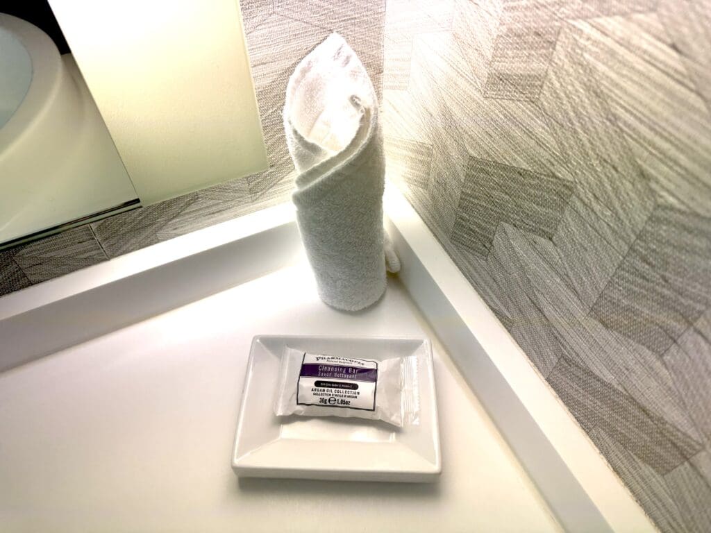 The image shows a bathroom countertop with a neatly rolled white towel standing upright. Next to it, there is a rectangular white dish holding a wrapped bar of cleansing soap. The background features a textured wall with a geometric pattern.
