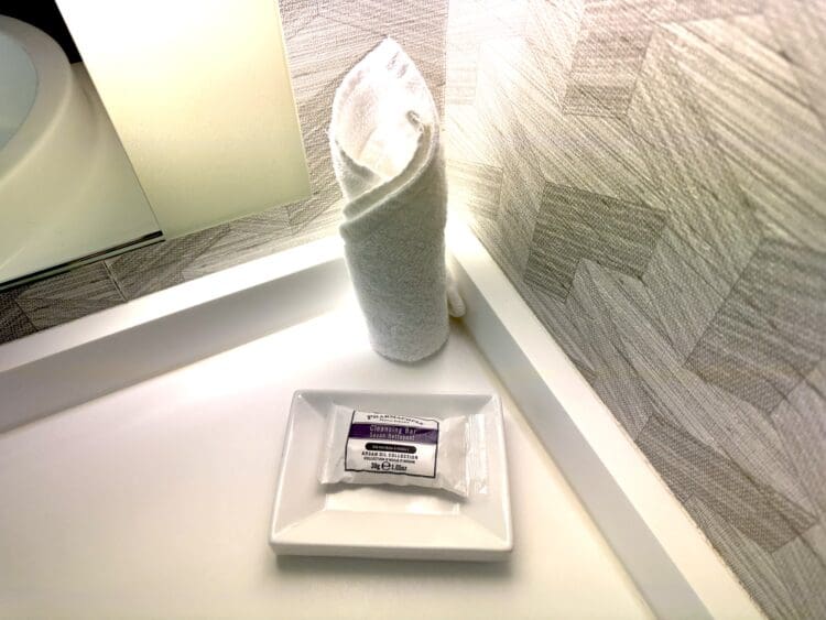The image shows a bathroom countertop with a neatly rolled white towel standing upright. Next to it, there is a rectangular white dish holding a wrapped bar of cleansing soap. The background features a textured wall with a geometric pattern.