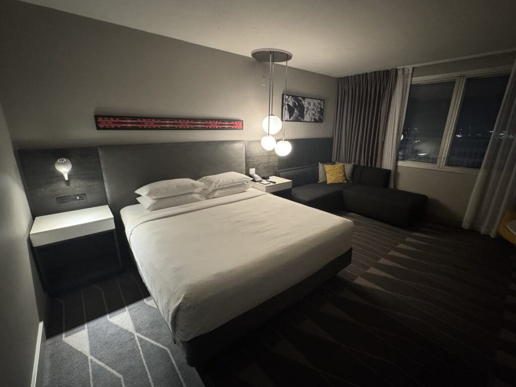 The image shows a modern hotel room with a large bed featuring white linens and pillows. There is a nightstand with a lamp on the left side of the bed. Above the bed, a piece of abstract art is mounted on the wall. To the right, there is a dark-colored sofa with a yellow cushion, and a window with closed curtains. A set of hanging pendant lights is positioned above the nightstand. The room has a striped carpet and a neutral color scheme.