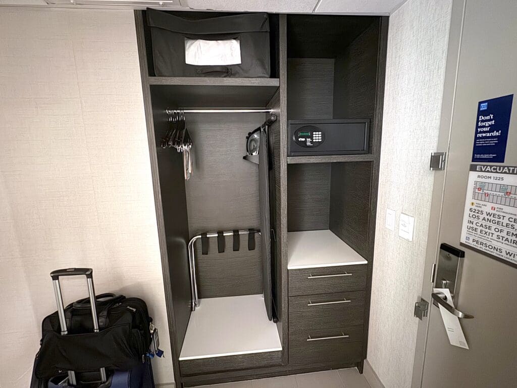 The image shows a hotel room closet area. Inside the closet, there are several hangers on a rod, an iron, and an ironing board. Above, there is a shelf with a bag or storage box. To the right, there is a safe on a shelf. Below the safe, there are two drawers. In front of the closet, there is a suitcase with a smaller bag on top. On the door, there is an evacuation plan and a sign about rewards.