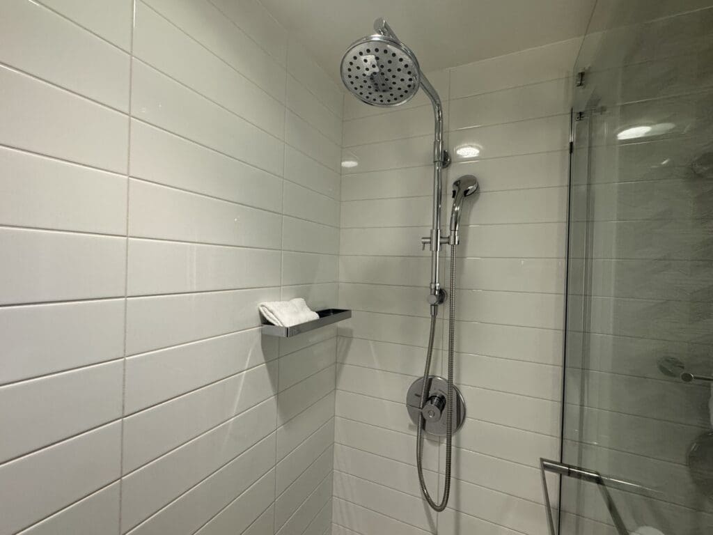The image shows a modern shower area with white tiled walls. There is a chrome showerhead and a handheld shower attachment. A small shelf holds a folded white towel. A glass door is partially visible on the right side.