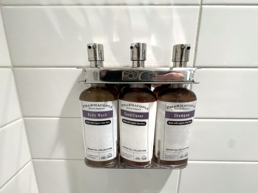 The image shows three bottles mounted on a wall in a bathroom setting. The bottles are labeled as "Body Wash," "Conditioner," and "Shampoo," all from the "Pharmacopia Natural Bodycare" brand. They are part of the "Argan Oil Collection" and are noted to be made with organic Aloe Vera, cruelty-free, and vegan. The bottles are held in a metal holder attached to white tiled walls.