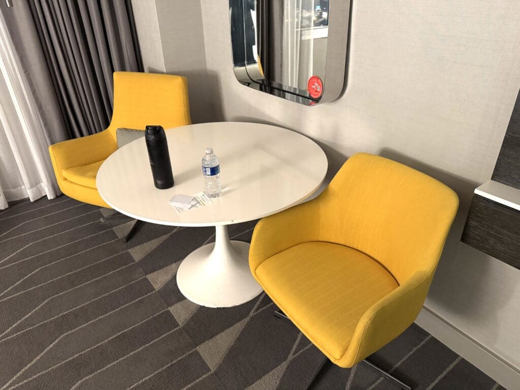 The image shows a small, modern room setup with a round white table and two yellow chairs. On the table, there is a black thermos, a plastic water bottle, and some papers. The floor has a gray carpet with a striped pattern, and there are gray curtains in the background. A mirror is mounted on the wall above the table.