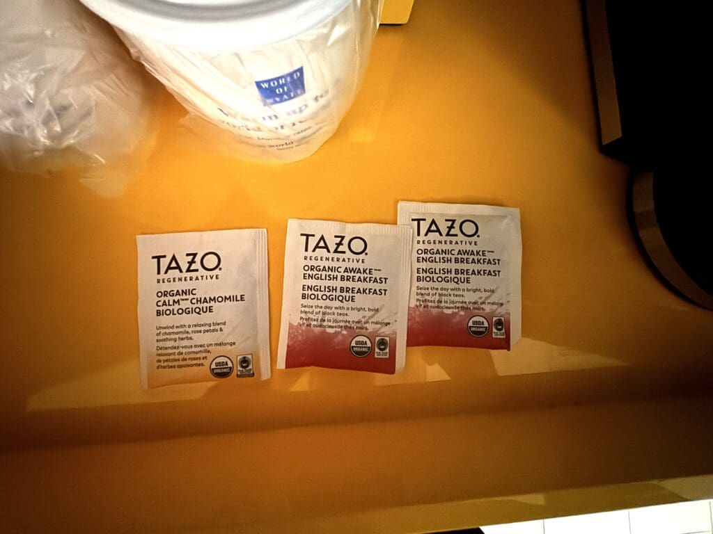 The image shows three packets of Tazo tea on a yellow surface. The packets include two "Organic Awake English Breakfast" and one "Organic Calm Chamomile." There is also a white cup with a blue label partially visible at the top left corner.