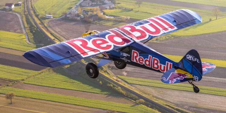 A small aircraft with "Red Bull" branding on its wings and body is flying over a patchwork of green and brown fields. The landscape below includes a few houses and winding roads. The plane is captured in mid-flight, showcasing its vibrant blue and red colors.