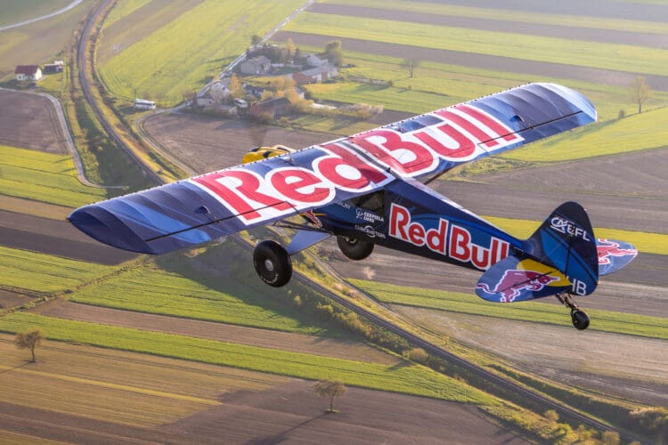 A small aircraft with "Red Bull" branding on its wings and body is flying over a patchwork of green and brown fields. The landscape below includes a few houses and winding roads. The plane is captured in mid-flight, showcasing its vibrant blue and red colors.