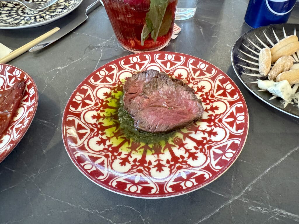 A slice of medium-rare beef is placed on a decorative red and white plate with a green sauce underneath. The plate is on a gray marble surface. Nearby, there are other plates with food, including a plate with small pastries and cheese, and a glass with a red drink and mint leaves at the Capital One Landing at Washington National Airport