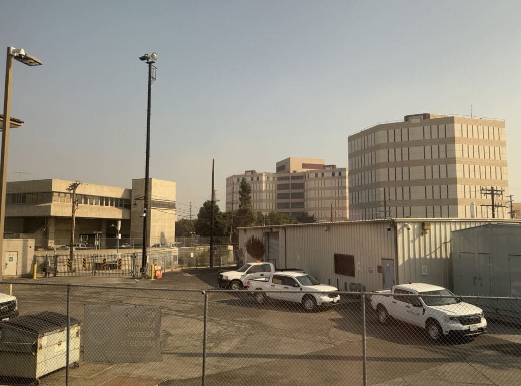 The image shows an industrial area with several large office buildings in the background. In the foreground, there is a fenced-off area with a few white utility vehicles parked on an asphalt surface. A metal building with a roll-up door is also visible. The sky is clear, and there are some trees near the buildings.