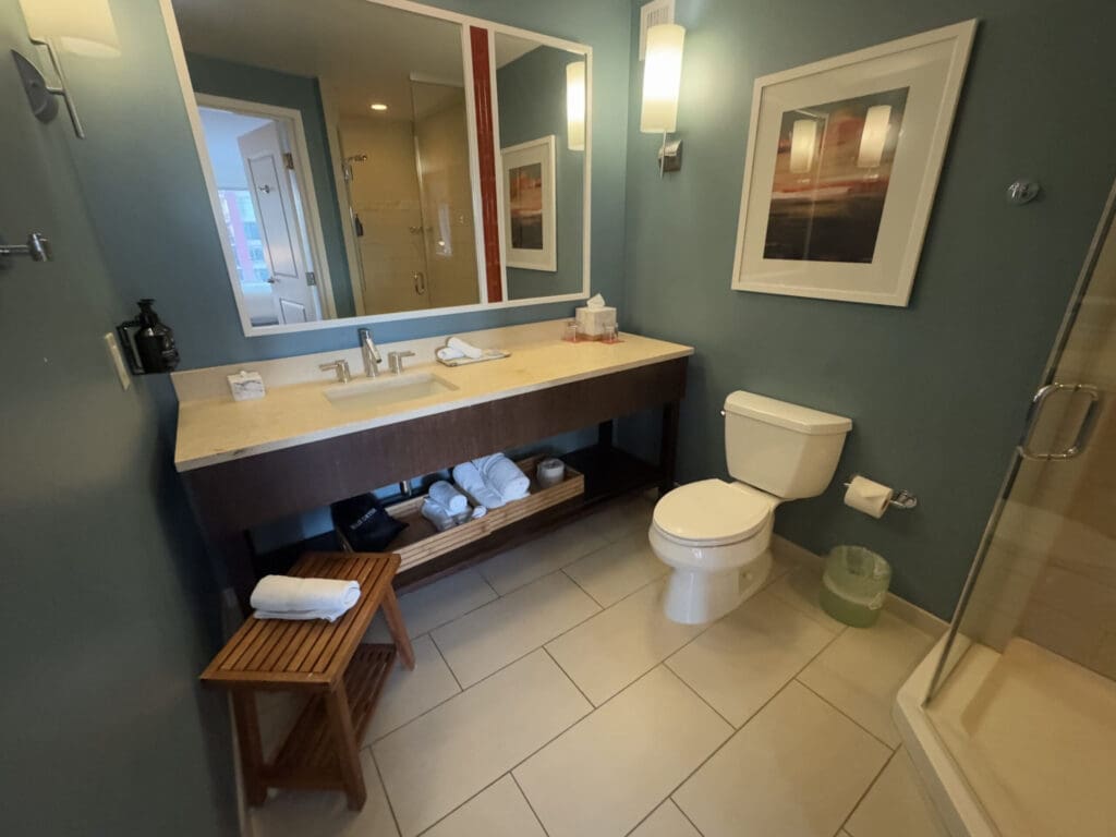 Hotel Indigo San Diego Gaslamp Quarter Studio Suite. The image shows a modern bathroom with teal walls. There is a large mirror above a beige countertop with a sink, faucet, and toiletries. Below the counter, there are shelves with towels and a black bag. To the right, there is a toilet with a roll of toilet paper on a holder and a small trash bin. A framed abstract artwork hangs on the wall. On the left, a wooden stool with a folded towel is placed on the tiled floor. A glass shower enclosure is partially visible on the right.