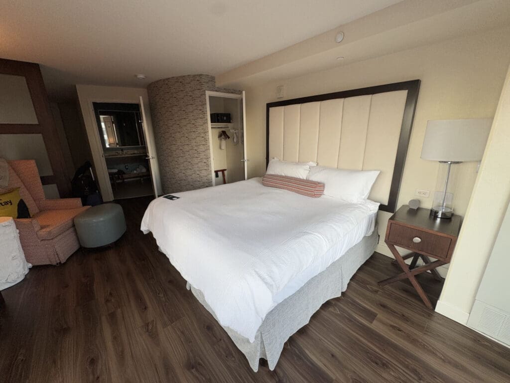 Hotel Indigo San Diego Gaslamp Quarter Studio Suite. The image shows a hotel room with a large bed featuring a padded headboard and white bedding. There is a striped bolster pillow on the bed. To the right of the bed is a wooden nightstand with a lamp. The room has wooden flooring, and in the corner, there is a pink armchair with a round ottoman. A closet with hangers and a safe is visible, and the bathroom can be seen through an open door.