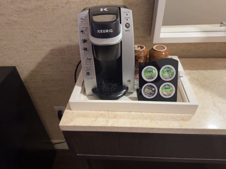 Hotel Indigo San Diego Gaslamp Quarter Studio Suite. The image shows a Keurig coffee maker placed on a countertop. Next to the coffee maker, there is a holder containing four coffee pods. Two brown ceramic mugs are also visible behind the coffee pods. The setup is on a tray, and the background includes a wall and a mirror.