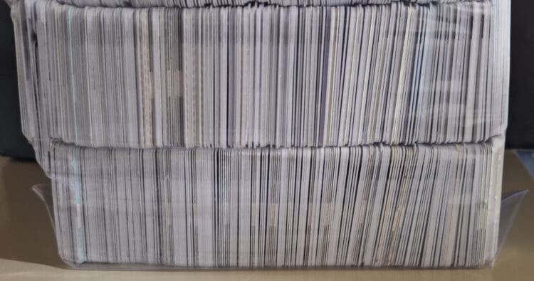 The image shows a large stack of cards, possibly trading or collectible cards, organized in two rows. The cards are tightly packed together and are encased in a clear plastic container. The edges of the cards are visible, showing a variety of colors and patterns.