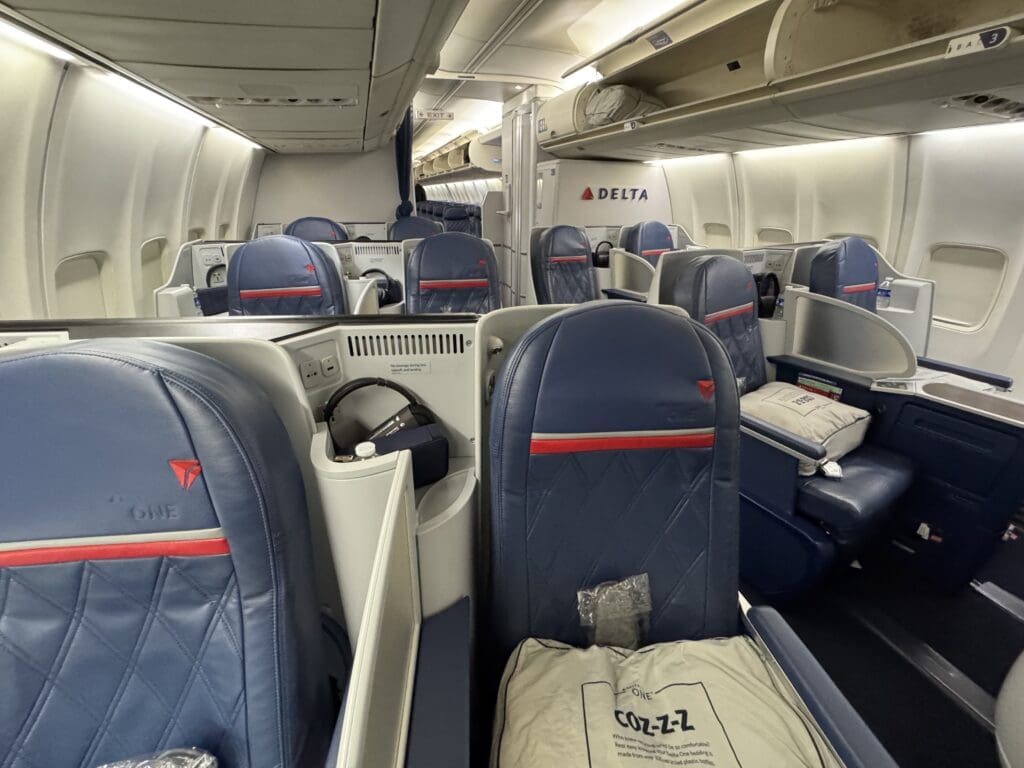 The image shows the interior of a Delta One business class cabin on an airplane. The seats are arranged in a spacious layout with blue leather upholstery and red accents. Each seat has a pillow and a blanket, and there are personal storage compartments and power outlets visible. The cabin is well-lit, and overhead bins are open with some luggage stored inside.